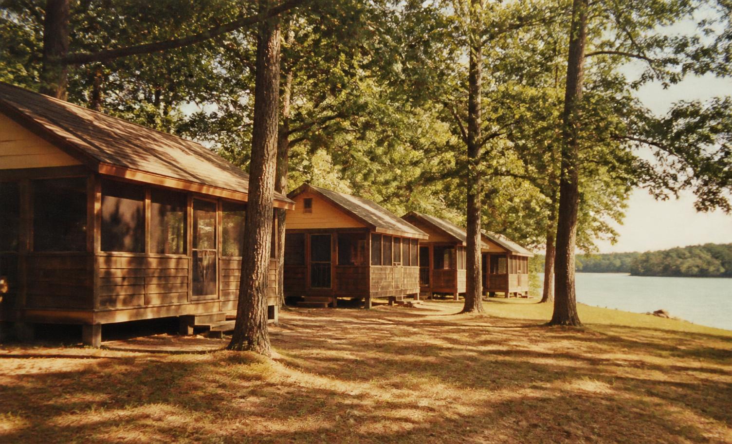 A row of wooden camp cabins under shade trees beside a lake